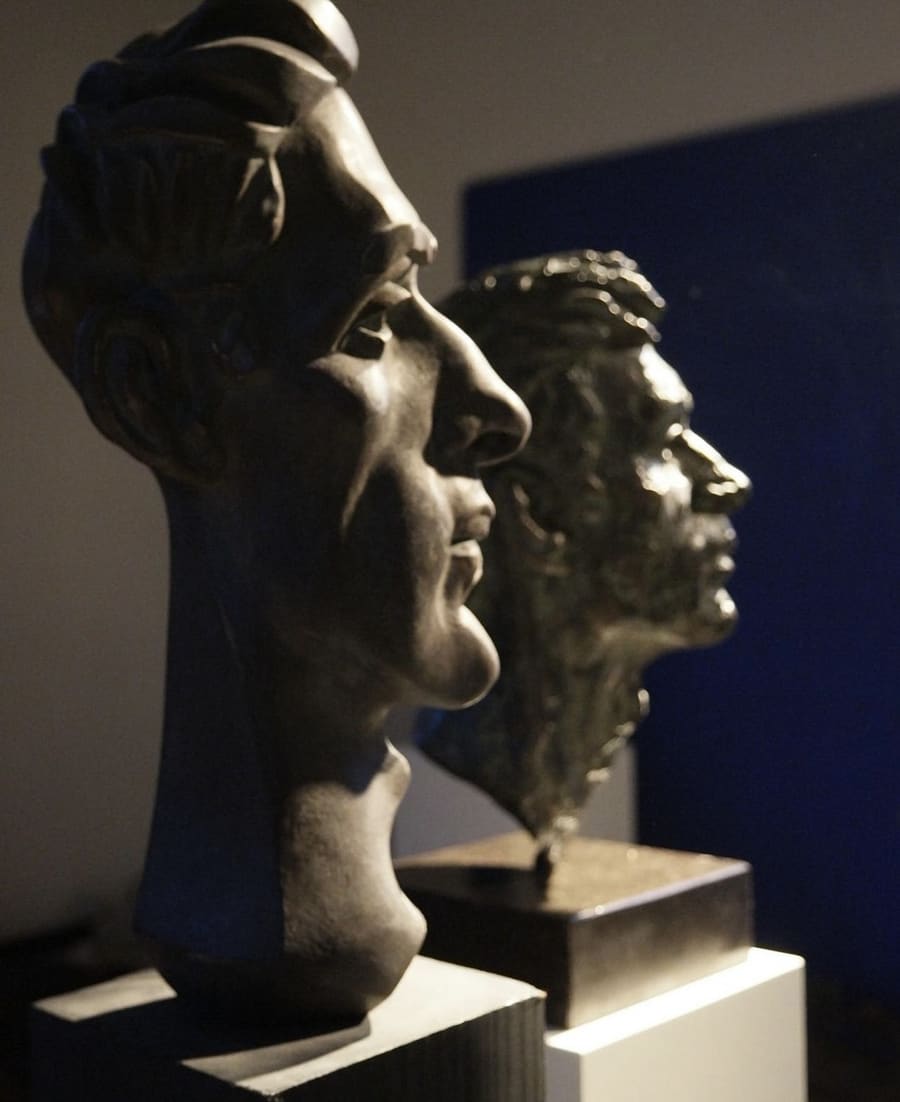 Twee bronzen portretbustes op sokkel, van opzij belicht.